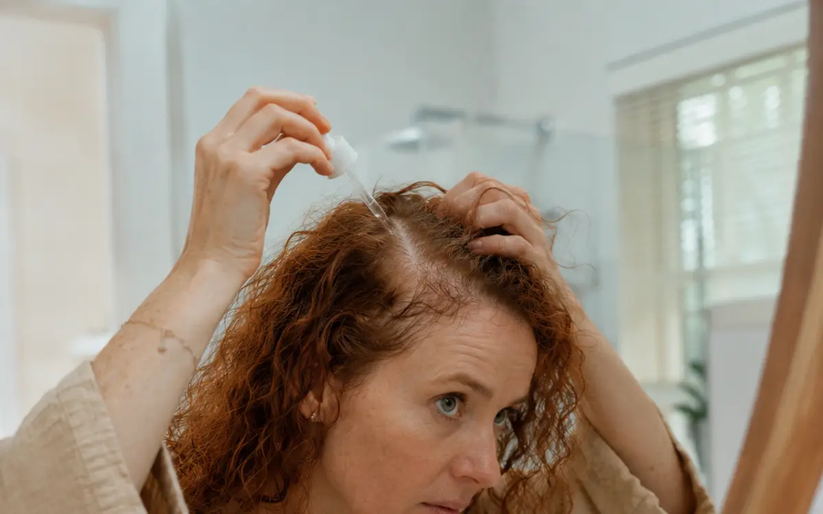 A woman applying a minoxidil dropper to her scalp at the hairline in front of a mirror